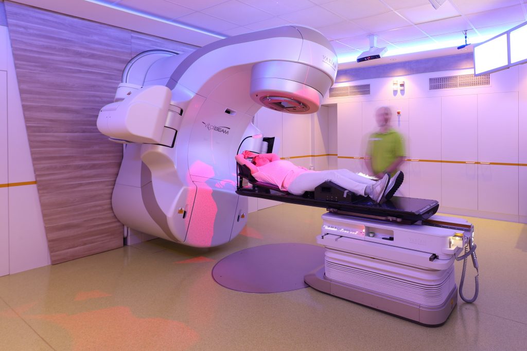 Patient liegt auf Behandlungsliege unter einem großen medizinischen Bestrahlungsgerät in einem Behandlungsraum